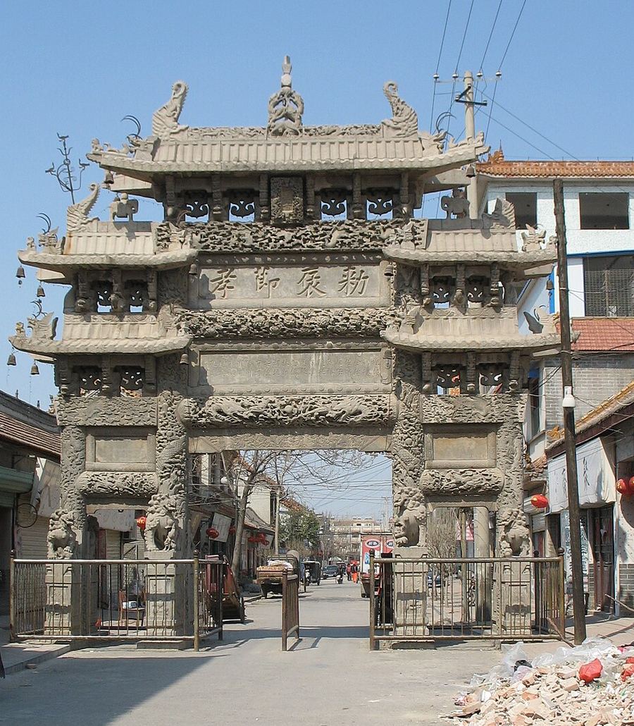 单县百寿坊正面，三间四柱式青石牌坊，檐下密布透雕，两侧为现代街市